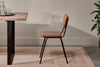 The nkuku Ukari Leather Dining Chair - Aged Tan, with its padded leather seat and black metal frame, sits by a rustic wooden table on a woven beige rug. Clear glasses top the table, with a textured off-white wall and green plant nearby.