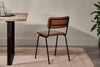 The Ukari Leather Dining Chair by nkuku in aged tan leather, featuring a padded seat and black metal frame, sits beside a rustic wooden table with clear glasses on a dark tray, atop a beige rug, with a potted plant and neutral wall.