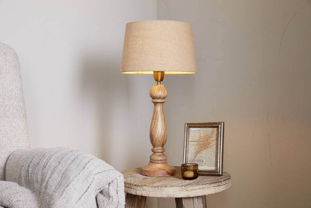 A cozy scene with a beige armchair, wooden side table, nkuku’s Usha Classic Wooden Table Lamp - Natural (eco-friendly mango wood), a candle, and a framed picture set against a neutral wall.