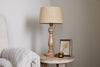 The nkuku Usha Classic Wooden Table Lamp - Natural, featuring a beige shade, rests on a rustic round table beside a small candle and framed photo. A light-textured armchair appears on the left against a plain, light wall.