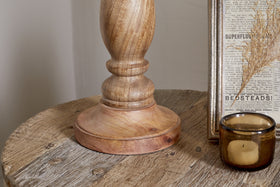 A close-up of a round wooden table featuring the nkuku Usha Classic Wooden Table Lamp - Natural, a brown glass candle holder, and a framed vintage newspaper clipping with dried grass inside.