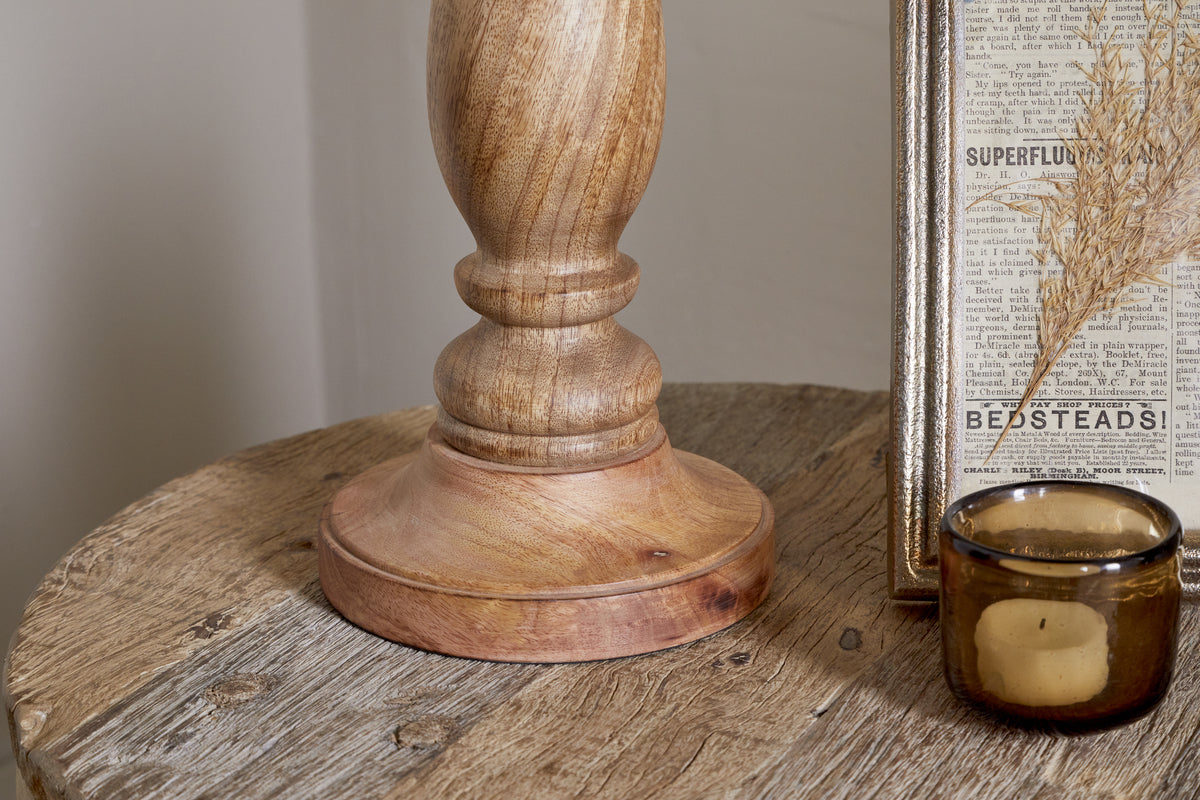 A close-up of a round wooden table featuring the nkuku Usha Classic Wooden Table Lamp - Natural, a brown glass candle holder, and a framed vintage newspaper clipping with dried grass inside.
