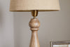 A close-up of the nkuku Usha Classic Wooden Table Lamp in natural mango wood with a beige fabric lampshade, placed beside a framed picture on a light-colored surface.