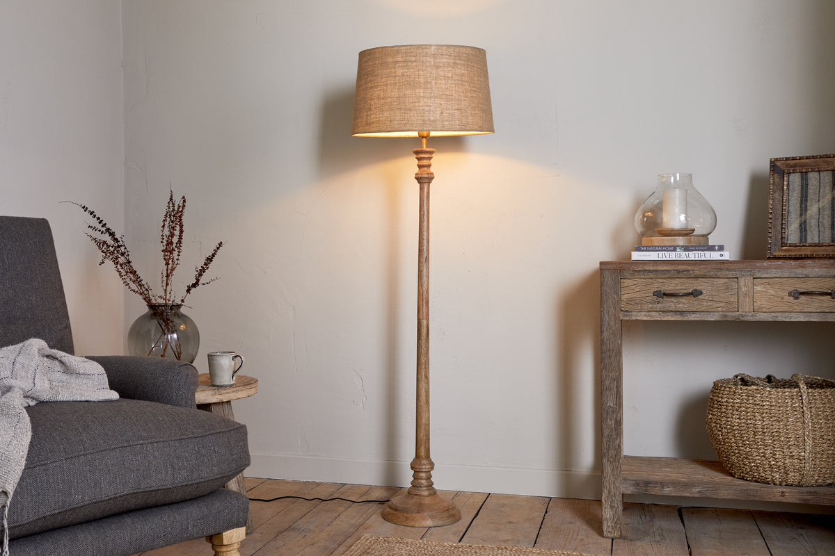 The nkuku Usha Classic Wooden Floor Lamp in natural stands lit beside a gray armchair with a knitted throw. To the right, a rustic wooden table displays a glass vase, books, and basket beneath, enhancing the cozy neutral vibe.