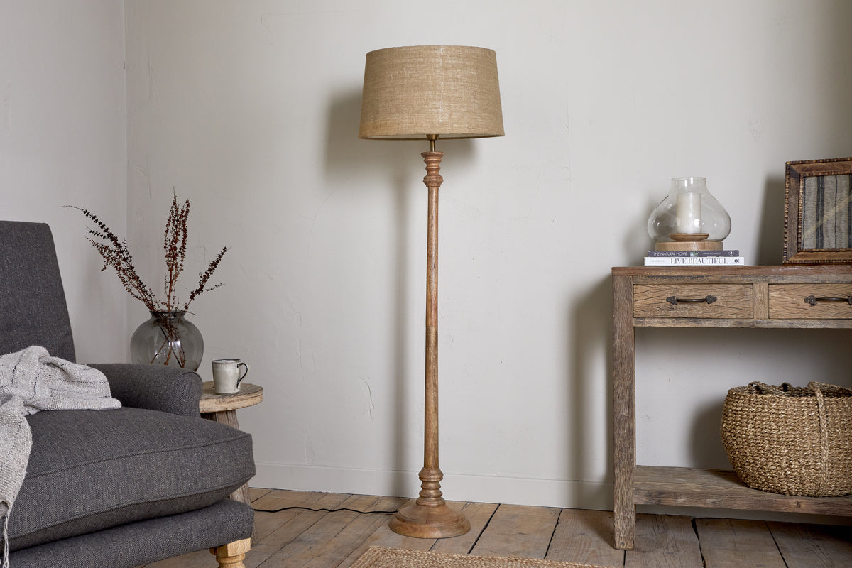 A cozy living room showcases the nkuku Usha Classic Wooden Floor Lamp - Natural, with a beige shade, positioned between a gray armchair with a draped blanket and a rustic wooden console table decorated with a basket and dried flowers.