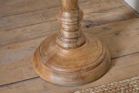 A close-up of the Usha Classic Wooden Floor Lamp - Natural by nkuku shows its round, carved mango wood base with a natural finish and visible grain on a rustic floor, alongside a tan woven rug in the corner.