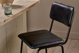 The nkuku Ukari Counter Chair in aged black leather with metal legs stands by a rustic wooden table. On the table are a clear ribbed glass and a round light brown item, set against light wood floors and neutral beige walls.
