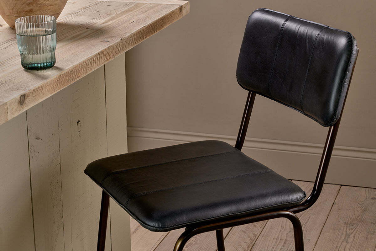 The nkuku Ukari Counter Chair in aged black leather with metal legs stands by a rustic wooden table. On the table are a clear ribbed glass and a round light brown item, set against light wood floors and neutral beige walls.