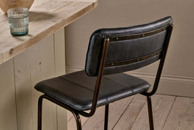 The nkuku Ukari Counter Chair in Aged Black, made with buffalo leather and a metal frame, sits by a light wood table. On the table is a clear ribbed glass of water. The floor features light wood planks and the wall is neutral beige.