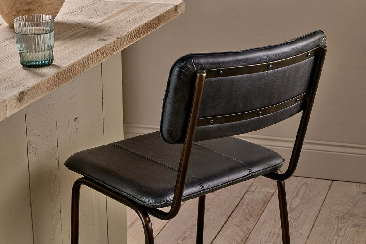 The nkuku Ukari Counter Chair in Aged Black, made with buffalo leather and a metal frame, sits by a light wood table. On the table is a clear ribbed glass of water. The floor features light wood planks and the wall is neutral beige.