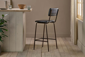 The nkuku Ukari Counter Chair - Aged Black, featuring a padded seat and backrest, stands on light wood flooring by a pale kitchen island with bowls and glasses as sunlight filters through sheer curtains near a green plant.
