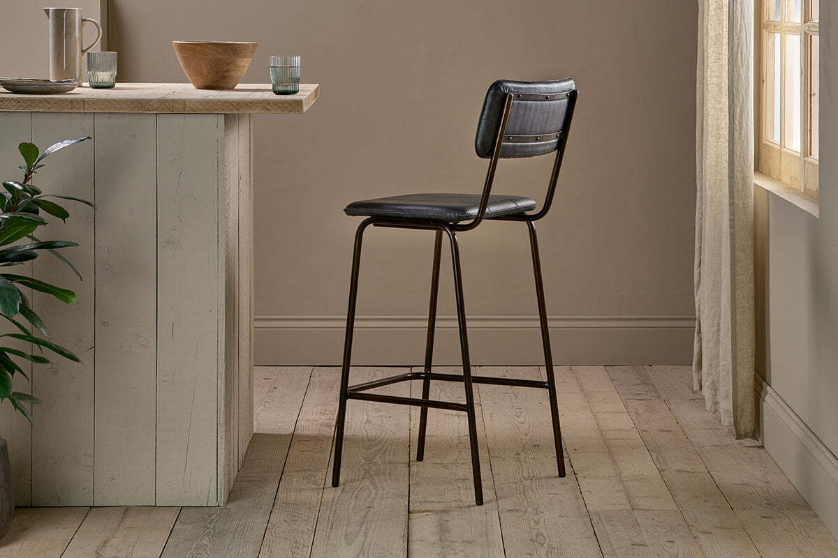 The nkuku Ukari Counter Chair - Aged Black, featuring a padded seat and backrest, stands on light wood flooring by a pale kitchen island with bowls and glasses as sunlight filters through sheer curtains near a green plant.