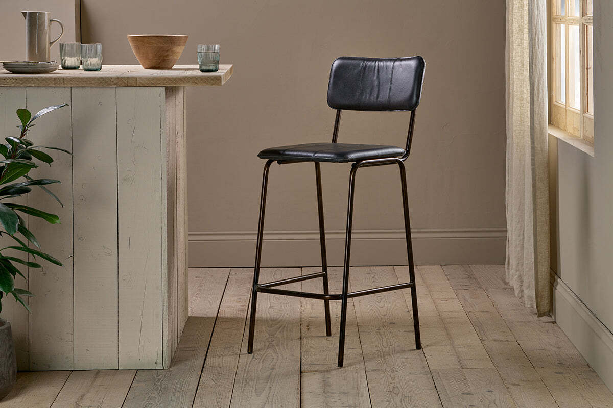 The nkuku Ukari Counter Chair in aged black leather and metal stands on light wooden flooring next to a rustic, light kitchen island. Sunlight through sheer curtains brightens the minimal, neutral space with simple decor.