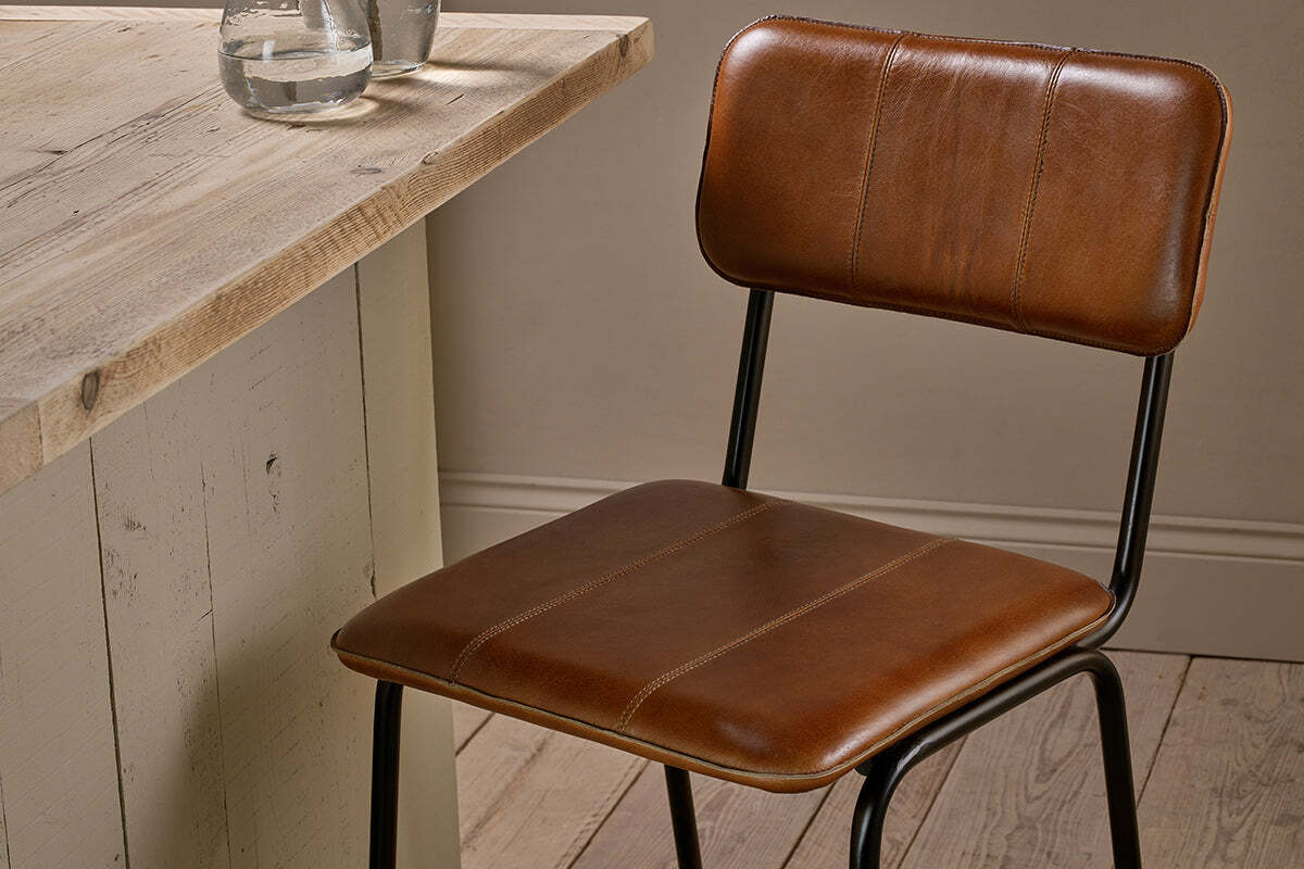 The Ukari Counter Chair - Aged Tan by nkuku, featuring brown handmade buffalo leather, black metal legs, and a rectangular cushioned backrest, sits by a rustic wooden table for a warm, minimalistic look.