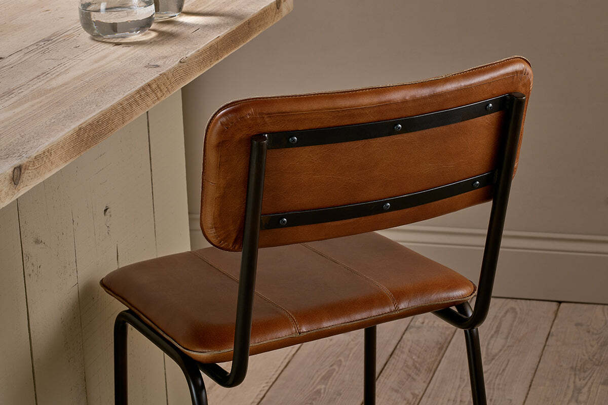 The nkuku Ukari Counter Chair - Aged Tan, featuring a handmade buffalo leather padded seat and backrest, stands by a rustic wooden table with a beige base. Two glasses of water sit on the table over light wood floors.