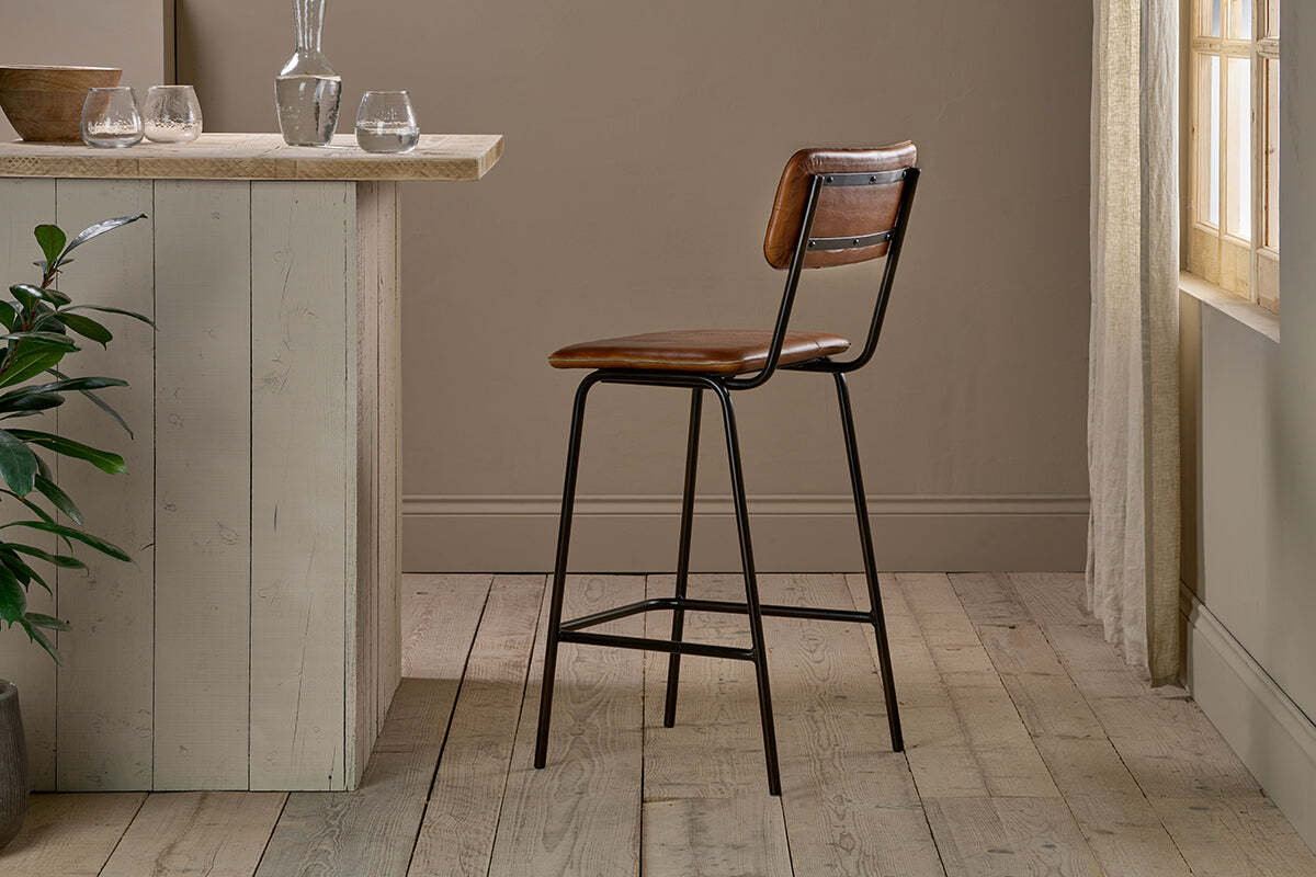 The nkuku Ukari Counter Chair - Aged Tan, with buffalo leather and a metal frame, is set beside a light wood kitchen island in a sunlit room with light wood floors, neutral walls, glassware, a plant, and sheer curtains.