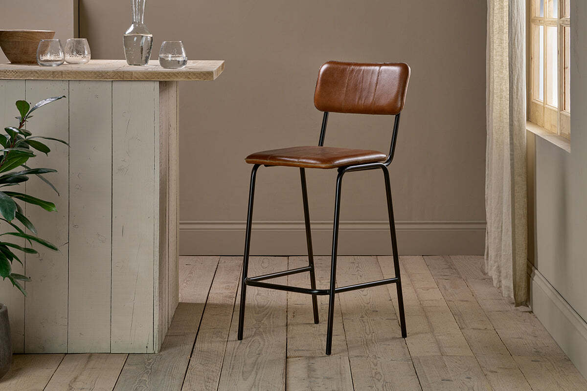 The nkuku Ukari Counter Chair - Aged Tan features a padded seat and back, slender black metal legs with footrest, standing by a rustic wooden counter in a neutral room with wood flooring and sunlight streaming in.