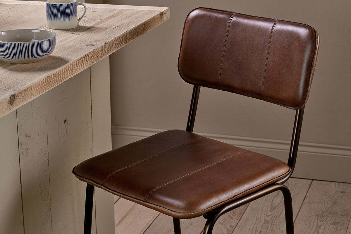 The nkuku Ukari Counter Chair in chocolate brown stands by a rustic wooden table. Two mugs and a patterned bowl sit atop the table, adding to the cozy, minimalist vibe of the wood-floored, neutral-walled space.