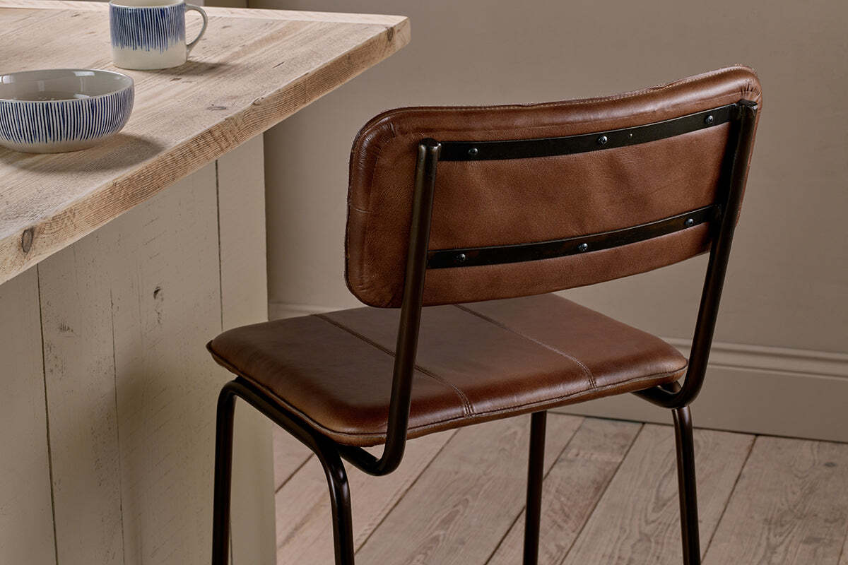 The Ukari Counter Chair - Chocolate Brown by nkuku, featuring black metal legs, is placed at a light wooden counter with blue and white patterned bowls and mugs, set against wood floors and neutral walls.