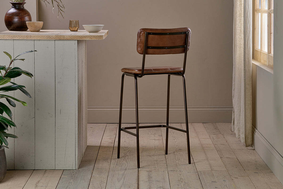 The nkuku Ukari Counter Chair in chocolate brown buffalo leather with slim black metal legs stands on light wood flooring by a pale wooden counter, accented by neutral bowls, a mug, and a plant as sunlight filters through a curtained window.