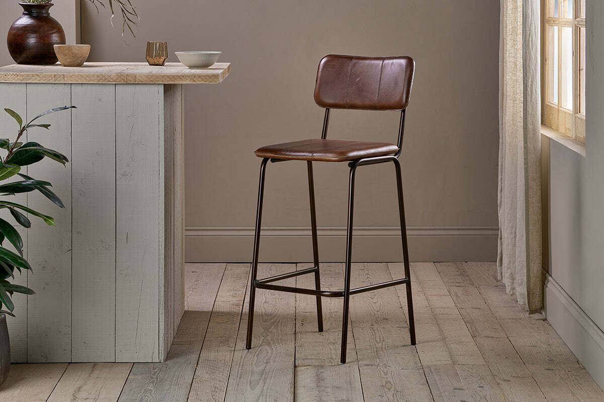 The nkuku Ukari Counter Chair - Chocolate Brown, featuring buffalo leather upholstery, stands on light wood flooring next to a pale gray counter with bowls, a cup, and a green plant beneath sunlit beige curtains.