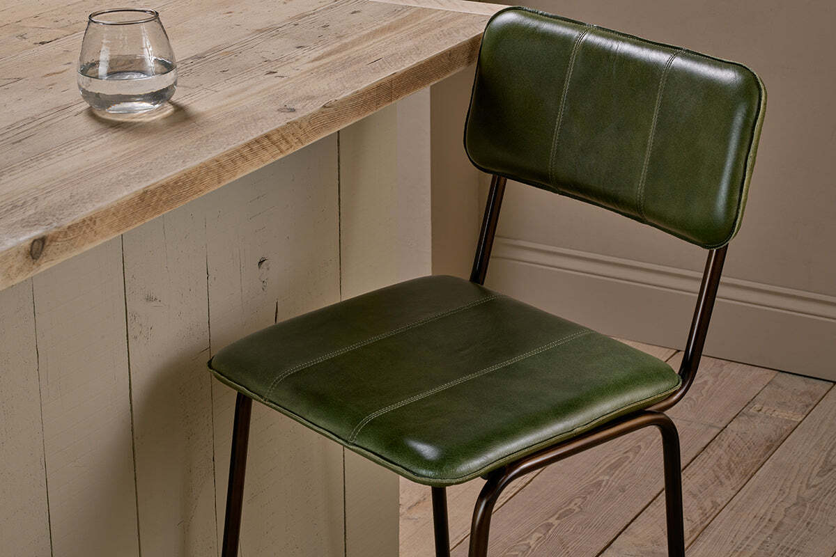 The nkuku Ukari Counter Chair in Rich Green, with a metal frame and leather upholstery, sits next to a rustic wood table. A round glass of water rests on the textured surface, surrounded by wooden floorboards and neutral walls.