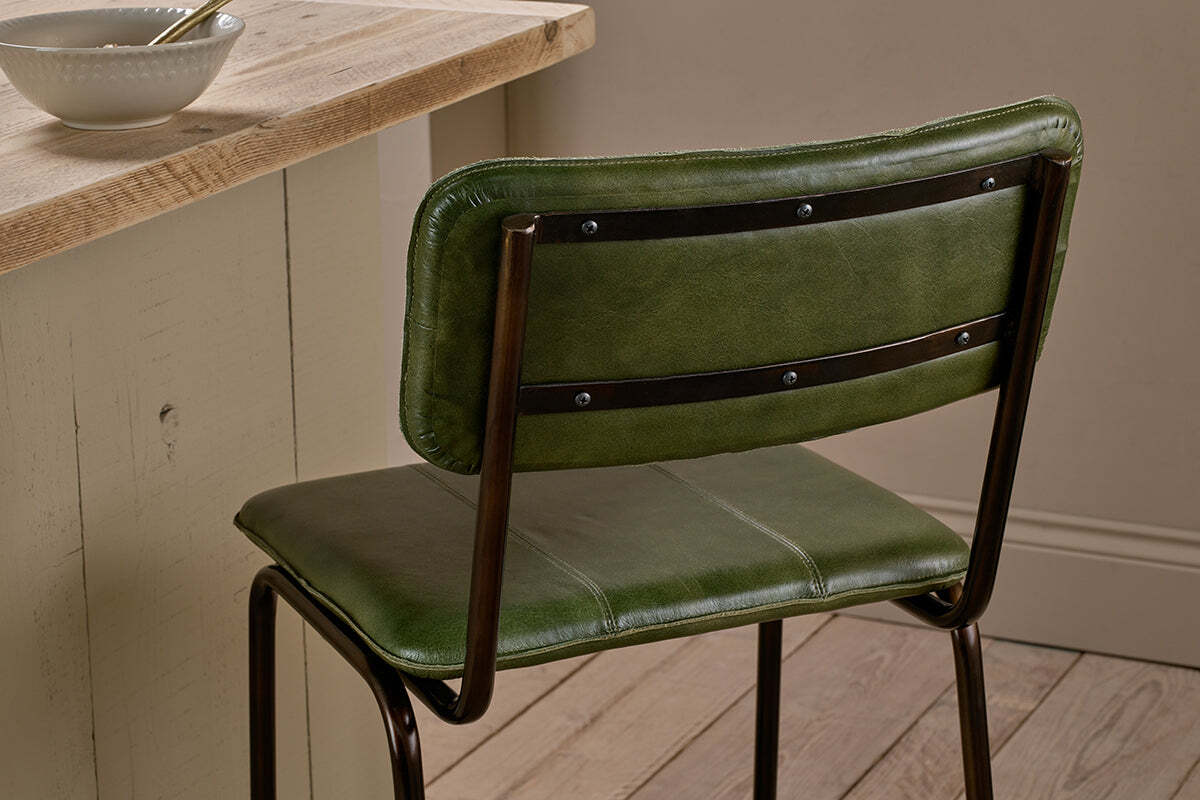A nkuku Ukari Counter Chair in rich green leather, featuring a padded seat and backrest, sits by a light wood counter. On the wooden floor nearby, a white bowl with spoon complements the room’s neutral walls.