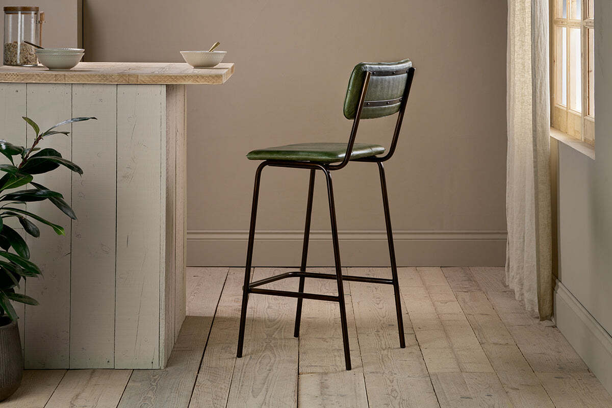 The nkuku Ukari Counter Chair - Rich Green, featuring a tall green leather seat and metal frame, stands by a light wood kitchen island in a bright room with wood floors, beige walls, a corner plant, and sunlight through sheer curtains.