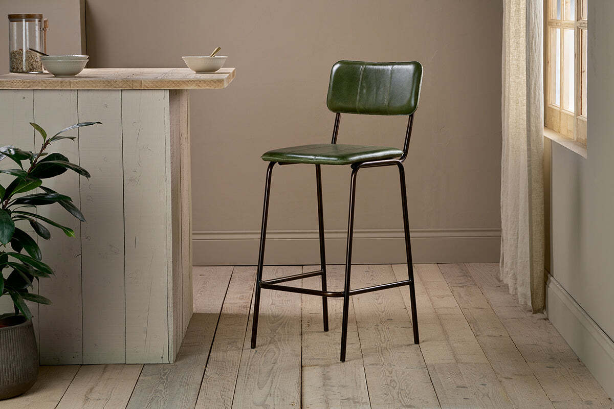The Ukari Counter Chair - Rich Green by nkuku, featuring a metal frame, stands on light wooden flooring next to a beige kitchen island with a potted plant and bowls, as soft sunlight streams through sheer curtains to the right.