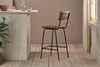 The Ukari Counter Chair - Aged Tan by nkuku, featuring a metal frame, stands on light wooden floors by a rustic pale wood counter topped with glass tumblers, a decanter, and small plants. Sunlight filters through sheer curtains from the right.