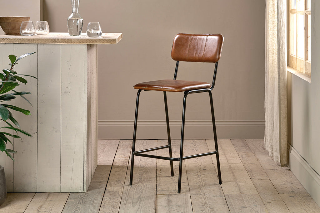 The nkuku Ukari Counter Chair in Aged Tan leather with a metal frame stands on a light wood floor by a rustic counter. Sunlight filters through sheer curtains, highlighting green plants and glassware arranged near the window.
