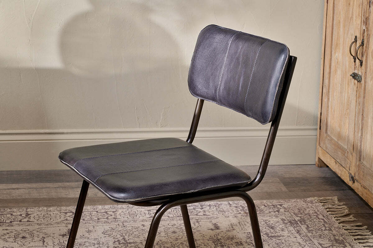 The Ukari Leather Dining Chair by nkuku, featuring an aged black padded buffalo leather seat and black metal frame, sits on a patterned rug beside a wooden cabinet against a beige wall with decorative trim and visible shadows.
