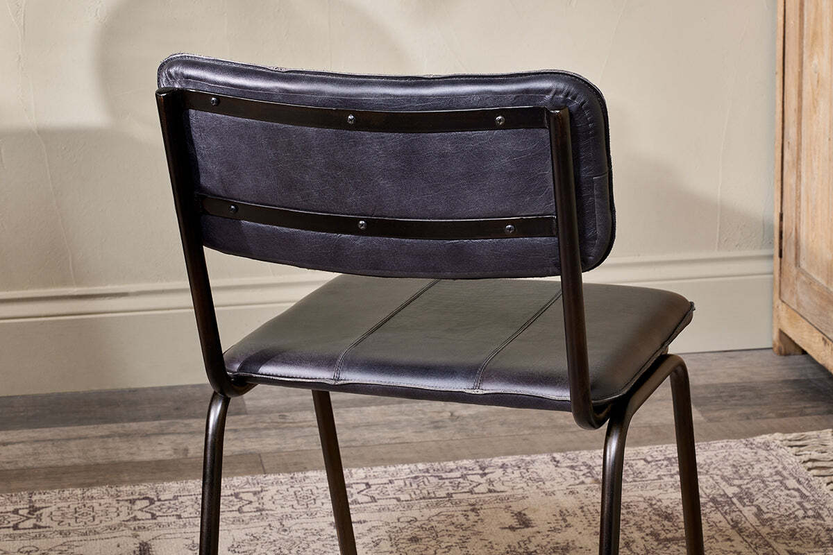 The Ukari Leather Dining Chair - Aged Black by nkuku is shown from behind, featuring a black metal frame and padded buffalo leather upholstery. It sits on a patterned rug over a wooden floor, with beige walls and wood furniture in the background.