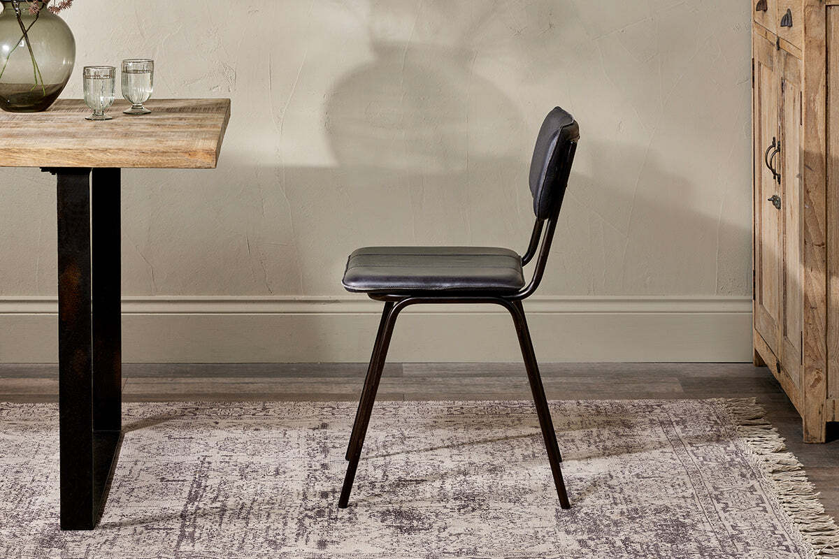 The nkuku Ukari Leather Dining Chair in Aged Black, featuring slim metal legs, sits sideways on a patterned rug next to a wooden table topped with two glasses and a vase. Behind it are a textured beige wall and part of a wooden cabinet.