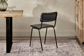 An nkuku Ukari Leather Dining Chair in Aged Black with buffalo leather upholstery sits on a patterned rug by a wooden table with glasses and a vase, set against light walls and near a wooden cabinet.