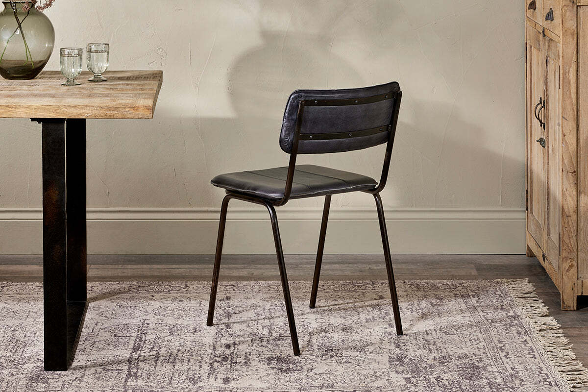 An nkuku Ukari Leather Dining Chair in Aged Black with buffalo leather upholstery sits on a patterned rug by a wooden table with glasses and a vase, set against light walls and near a wooden cabinet.