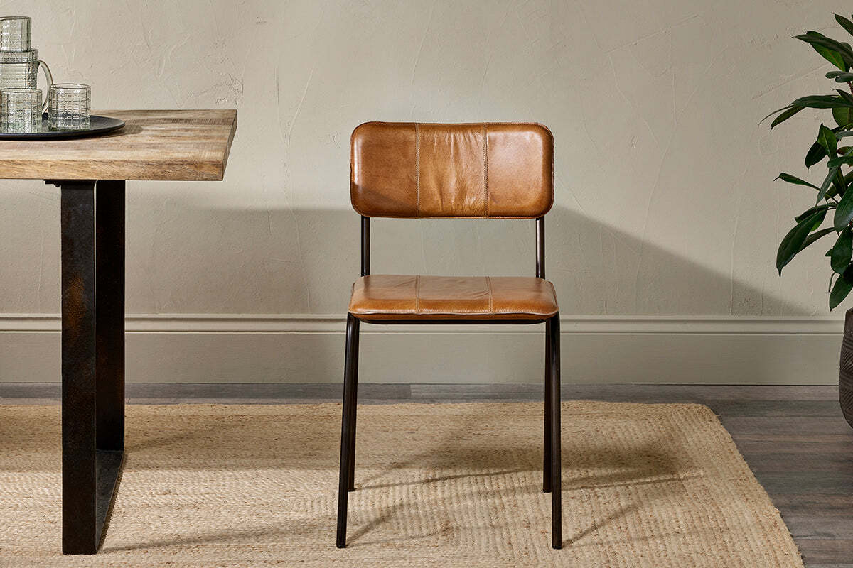The nkuku Ukari Leather Dining Chair in Aged Tan, featuring a metal frame, is placed on a jute rug beside a rustic wooden table.