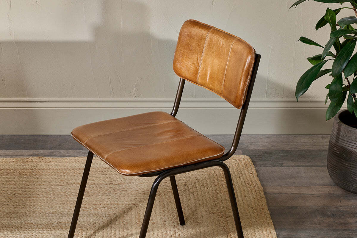 The nkuku Ukari Leather Dining Chair in aged tan, featuring padded buffalo leather upholstery and slim black metal legs, sits on a light woven rug atop a wood floor near a textured light wall, with a green potted plant partly visible to the right.