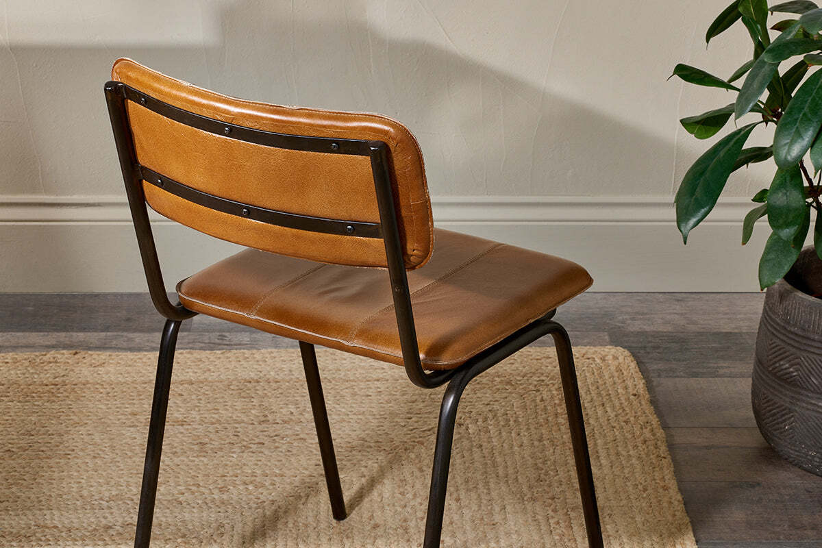 The nkuku Ukari Leather Dining Chair in aged tan, featuring a padded seat and backrest on a black metal frame, stands on a beige woven rug. A green potted plant is nearby, with a textured light wall in the background.