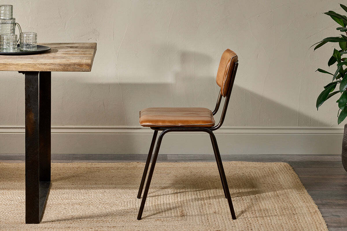 The nkuku Ukari Leather Dining Chair in Aged Tan with black metal legs sits on a woven beige rug by a wooden dining table. A leafy green plant adds brightness nearby, and glasses are neatly arranged on a tray atop the table.