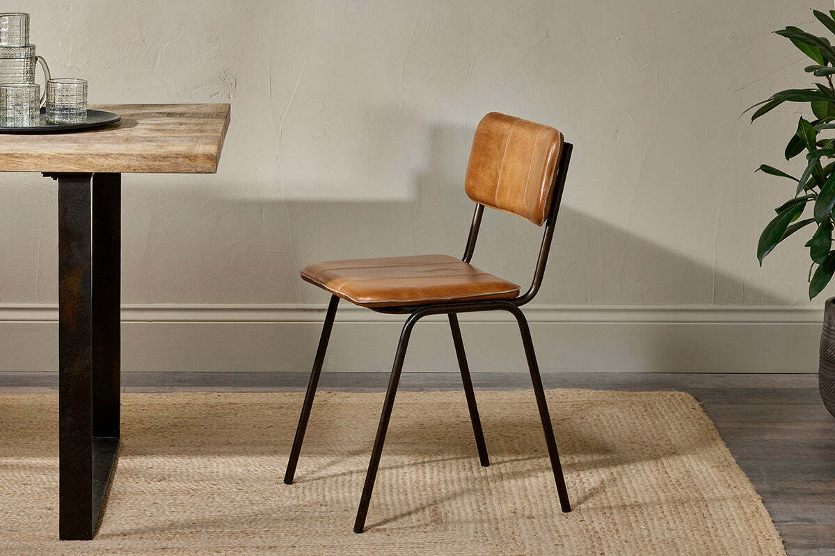 The nkuku Ukari Leather Dining Chair in Aged Tan, featuring cushioned leather upholstery and black metal legs, stands by a wooden table on a beige rug. A plant appears on the right against a neutral wall.