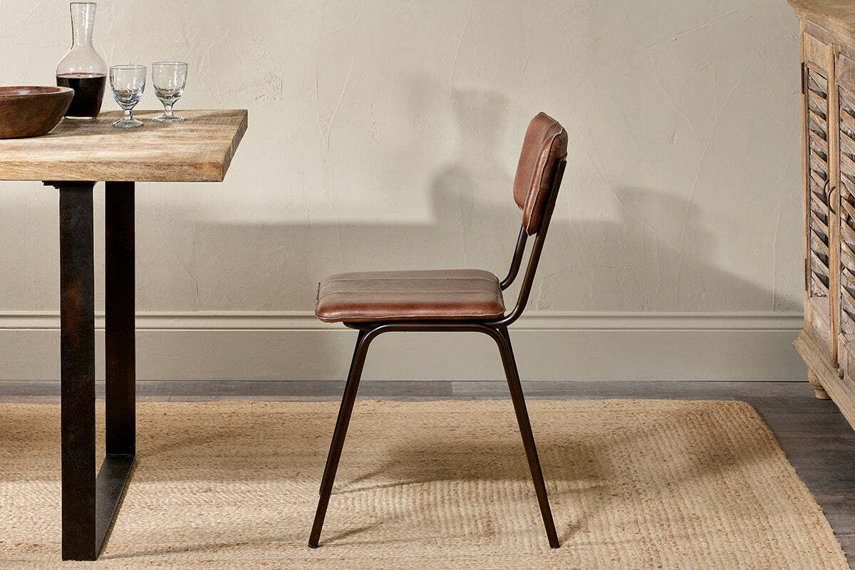 The nkuku Ukari Leather Dining Chair in chocolate brown, featuring a metal frame and padded leather seat, sits on a woven rug beside a wooden dining table topped with glasses, a decanter, and bowl, set by a textured beige wall and cabinet.