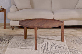 The Umar Mango Wood Coffee Table - Dark Brown by nkuku, featuring a circular design and three angular legs, is placed on a beige patterned rug in front of a light beige upholstered sofa. A wooden side table with a glass and book sits to the left.