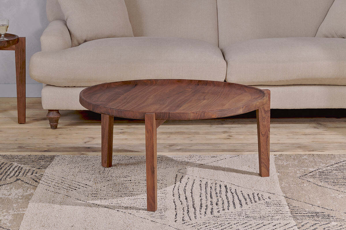 The Umar Mango Wood Coffee Table - Dark Brown by nkuku, featuring a circular design and three angular legs, is placed on a beige patterned rug in front of a light beige upholstered sofa. A wooden side table with a glass and book sits to the left.