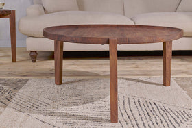 The Umar Mango Wood Coffee Table - Dark Brown by nkuku, featuring a sculptural circular design with three legs, is placed on a patterned beige rug in front of a light sofa, creating a cozy, modern living room setting.