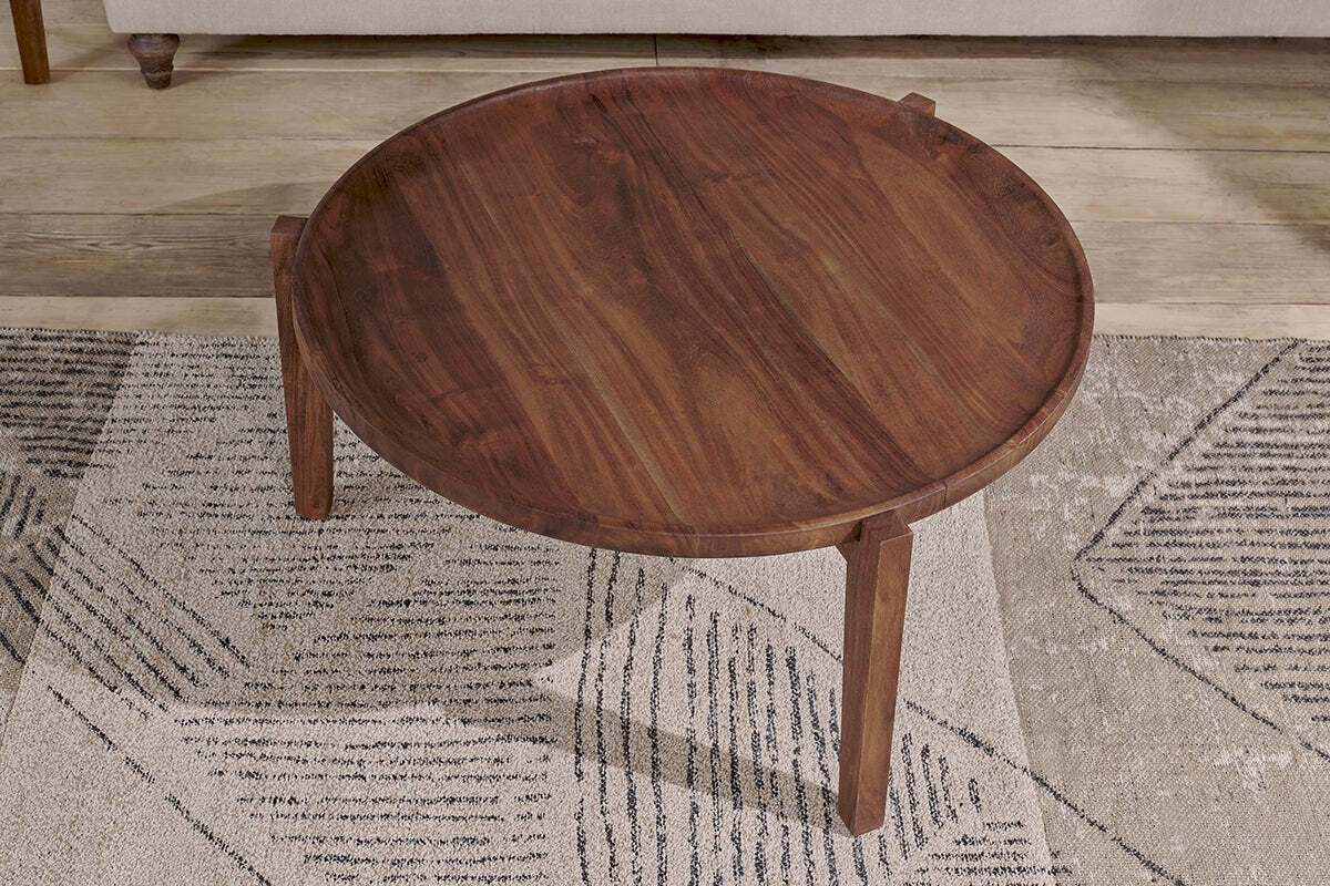 The nkuku Umar Mango Wood Coffee Table in dark brown, featuring a circular shape and tapered legs, sits on a rug with black and beige geometric patterns.