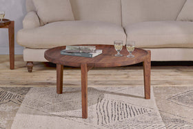 The nkuku Umar Mango Wood Coffee Table - Dark Brown, with its sculptural circular design, displays two glasses and books on a textured rug in front of a cream-colored sofa.