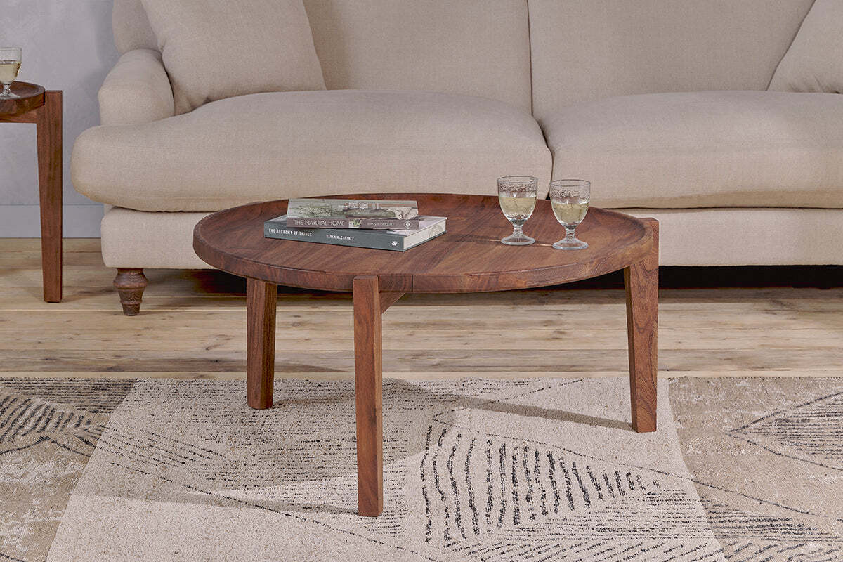 The nkuku Umar Mango Wood Coffee Table - Dark Brown, with its sculptural circular design, displays two glasses and books on a textured rug in front of a cream-colored sofa.
