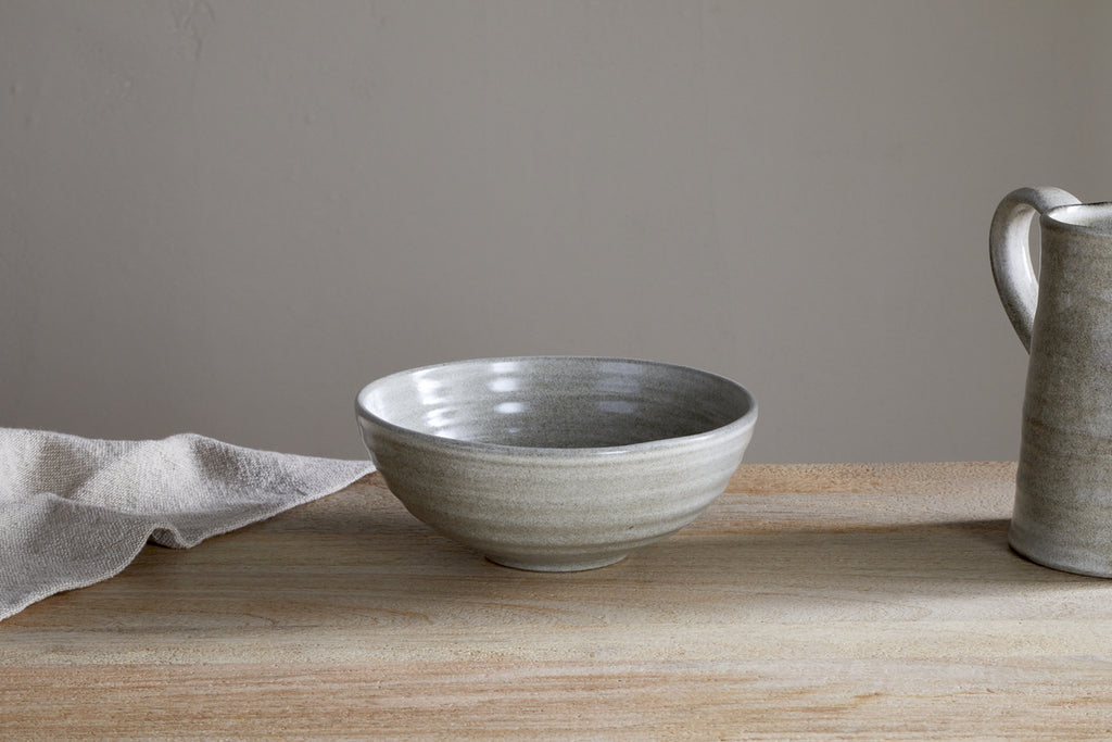 The Valinha Earthenware Cereal Bowl - Grey by nkuku features a light gray finish and textured spiral design. Shown on a wooden surface beside a beige cloth and matching pitcher, it’s set against a plain light-colored wall.