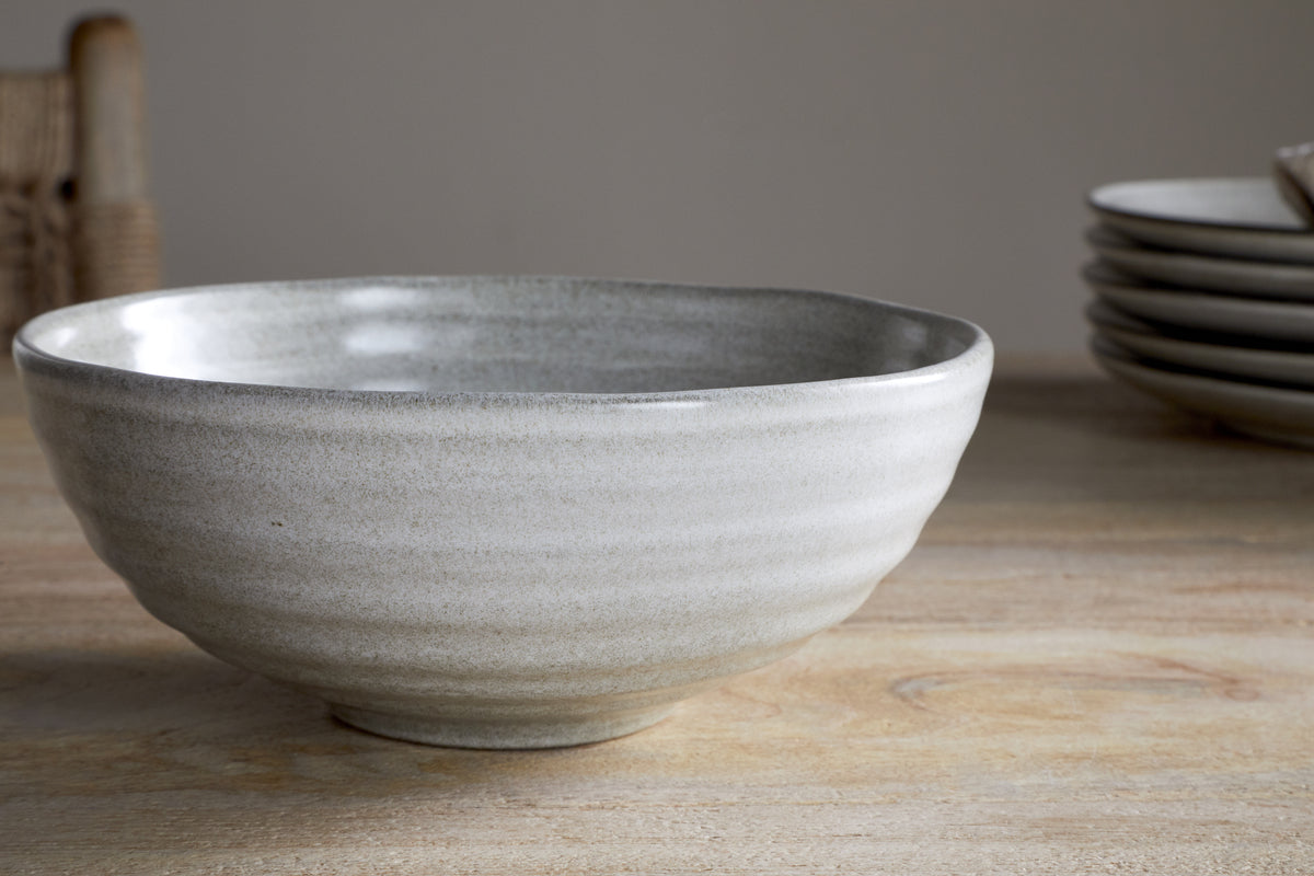 The Valinha Earthenware Cereal Bowl in grey by nkuku, featuring a textured spiral design, sits on a wooden table with matching plates blurred in the background and a woven chair partially visible to the left.